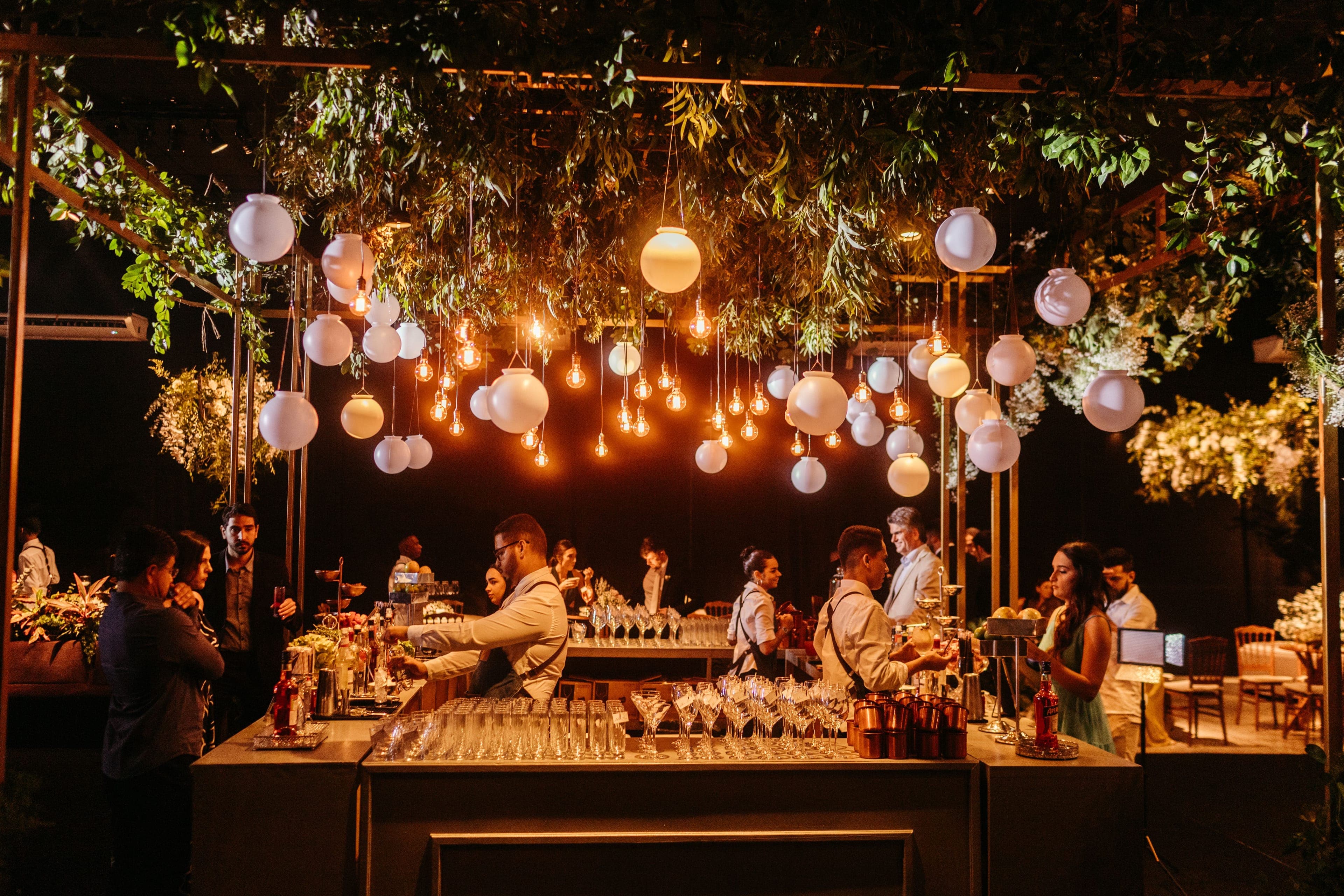 Barmen préparant des cocktails derrière un bar de mariage décoré de suspensions lumineuses et de verdure
