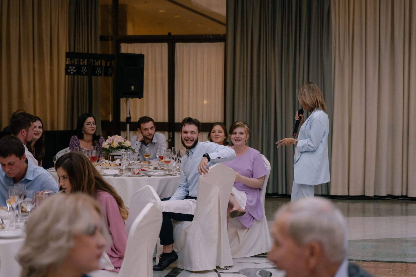 Invités attablés à des tables rondes nappées de blanc, écoutant un discours pendant une réception de mariage