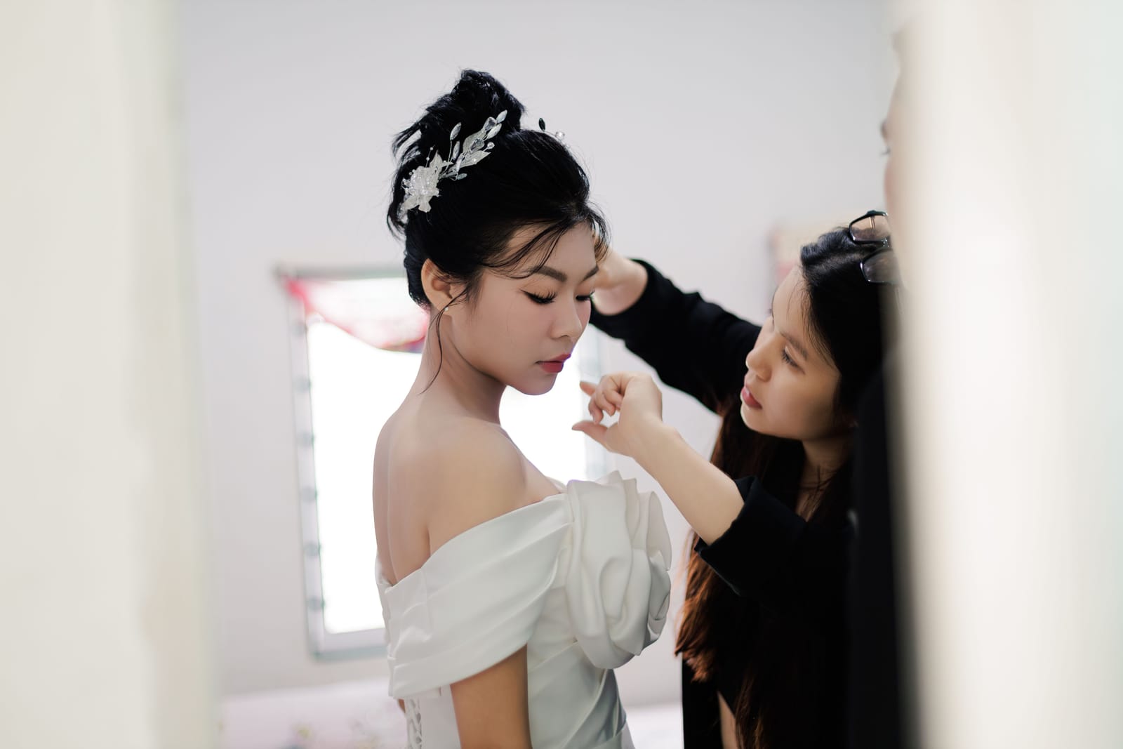 Mariée en robe blanche aux épaules dénudées, maquillée par une professionnelle dans une chambre lumineuse