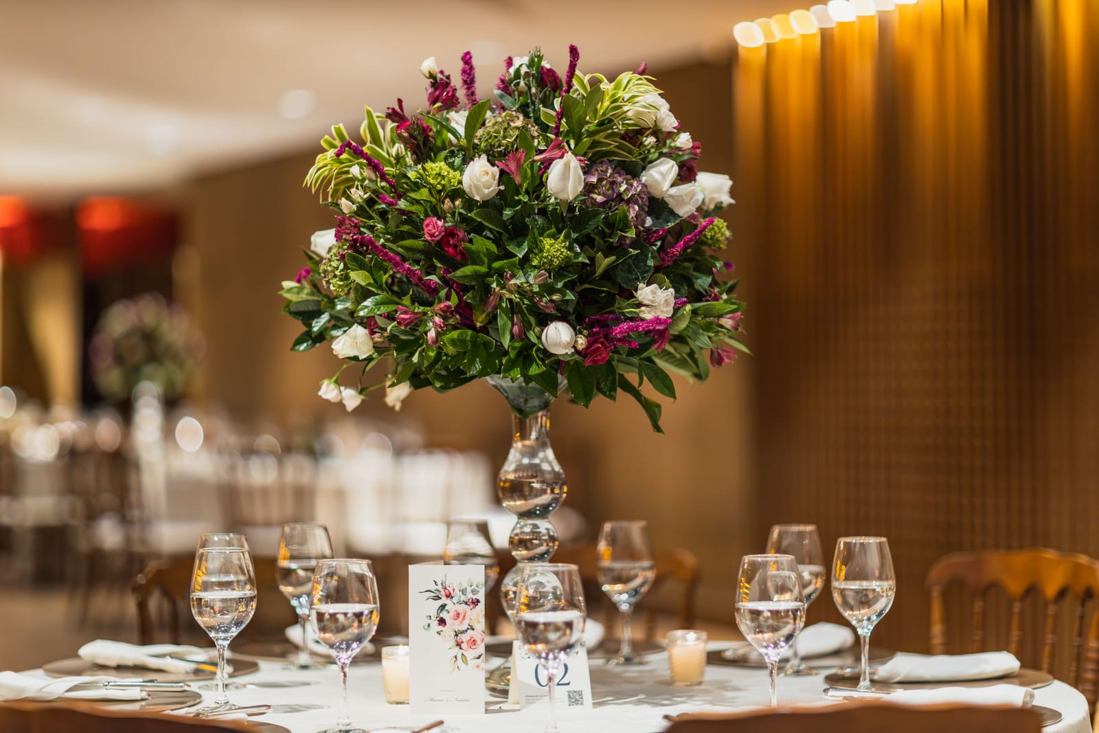 Centre de table haut sur pied de verre fin, roses et feuillages, sur une table de réception dressée