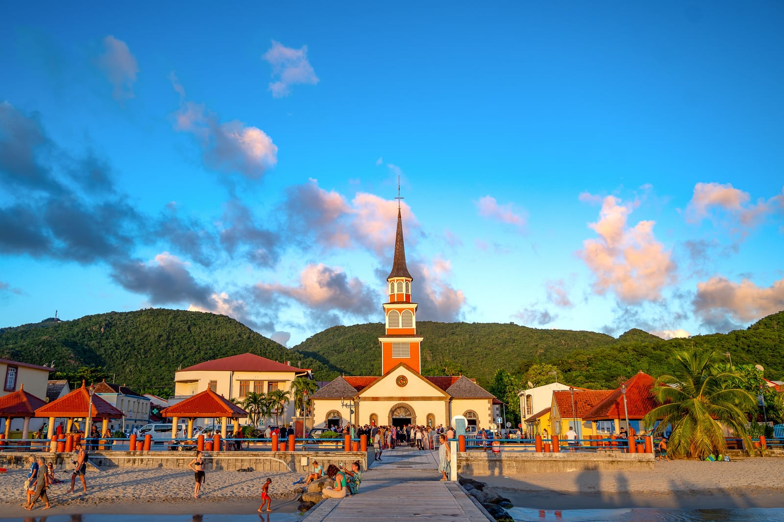 Église au clocher orange face au ponton et à la plage d'un bourg de Martinique, au pied des mornes