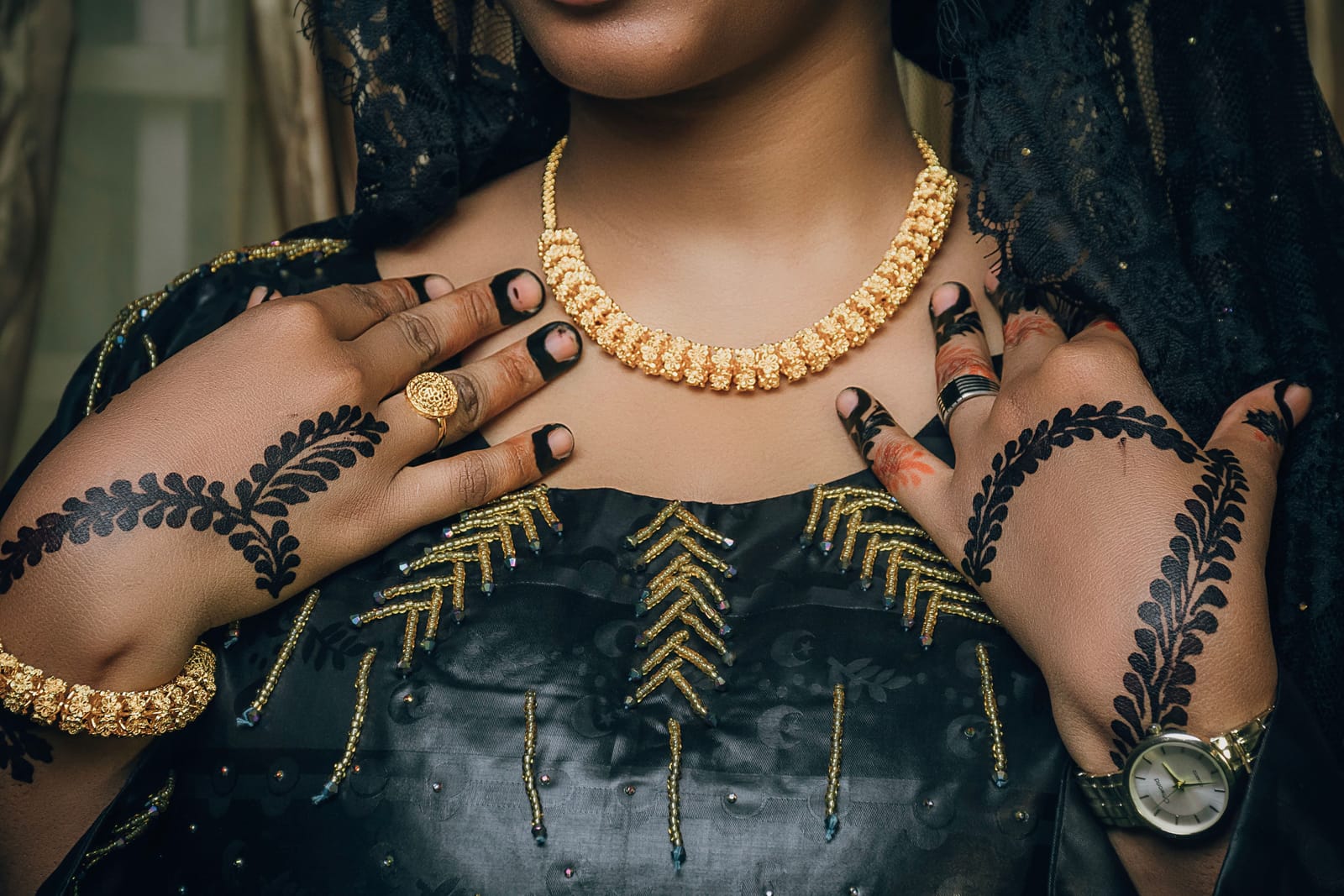 Mains de femme ornées de henné noir posées sur une tenue sombre brodée de perles dorées, avec un collier et un bracelet en or