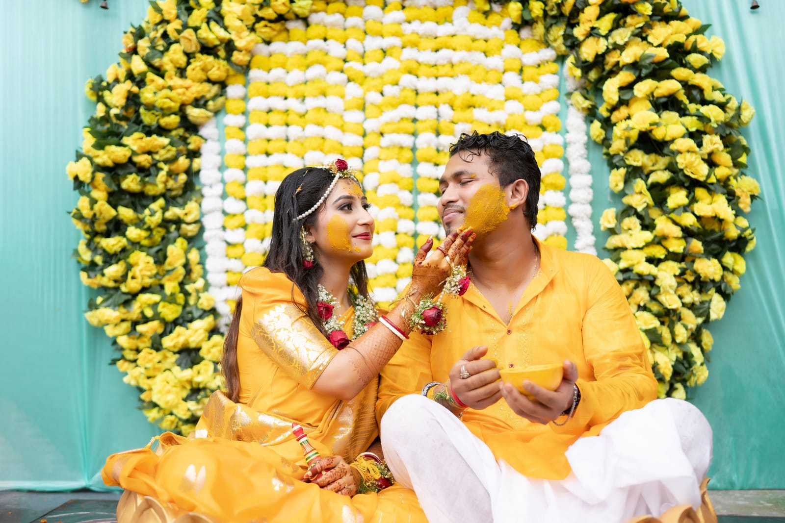 Pendant le haldi, la mariée applique de la pâte de curcuma sur la joue du marié devant un décor de fleurs jaunes et blanches