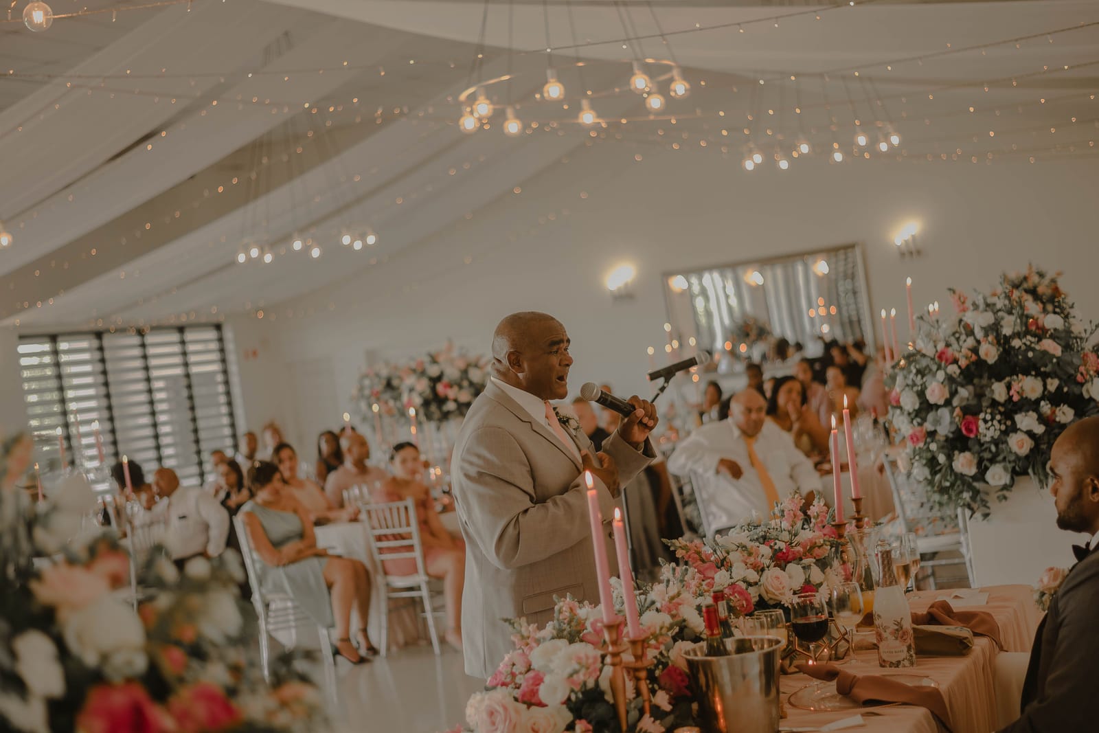 Un invité prononce un discours au micro pendant une réception de mariage, entouré de tables fleuries